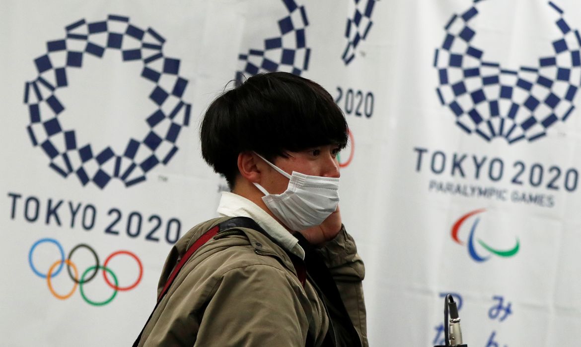 Homem com máscara de proteção em frente à logomarca dos Jogos Tóquio 2020
