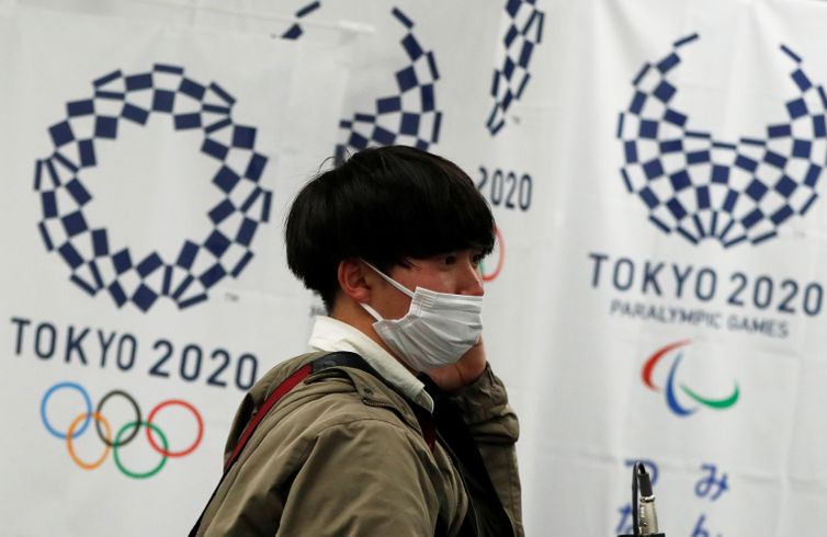 REUTERS/Issei Kato/Direitos Reservados Homem com máscara de proteção em frente à logomarca dos Jogos Tóquio 2020