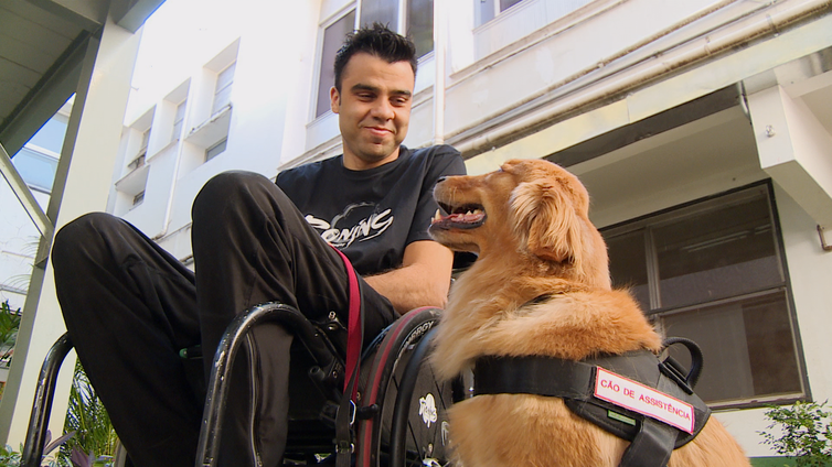 Descrição da foto: Em área externa, sentado na cadeira de rodas, o paratleta Lucas Junqueira olha para a cadela Paçoca. Sentada ao lado de Lucas, a cadela olha para ele.