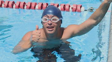 Dentro de uma piscina com raia, Gustavo, que tem síndrome de Down, está  de touca e de óculos de natação. De frente para nós, ele faz sinal de positivo, com o polegar estendido para cima