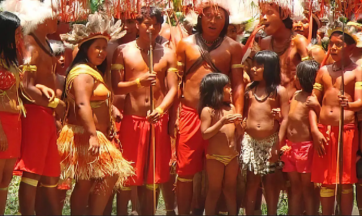 Imagem de um grupo indígena. Vestem roupas de penas e palhas ou tecido vermelho.