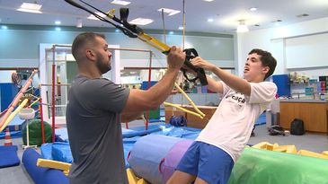 Sala de ginástica. Um rapaz com as mãos nos puxadores de uma fita de treinamento suspenso e o corpo inclinado para trás. De frente para ele, um educador físico com a mão direita na extremidade da fita.