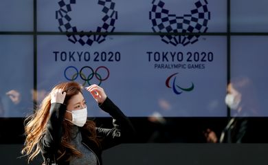 Mulher com máscara de proteção caminha em frente aos logos da Olimpíada e da Paralimpíada Tóquio-2020 / Jogos de Tóquio -anéis olimpícos, olimpíada, máscara, coi