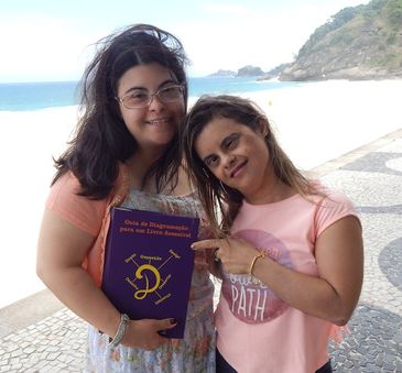 Fernanda Honorato e Fernanda Machado, jovens mulheres que têm síndrome de Down, estão em pé, lado a lado, no calçadão da praia. Fernanda Machado segura, sorridente, um livro, enquanto Fernanda Honorato aponta para a capa