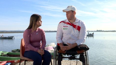 : Sentados lado a lado, Fernanda Honorato e Norberto Mourão, em uma cadeira de rodas. Ao fundo, barcos em uma lagoa