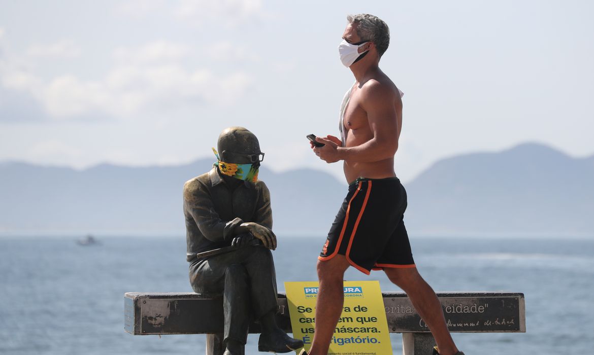 Um homem caminha ao lado da estátua do escritor brasileiro Carlos Drummond de Andrade que usa máscara protetora no primeiro dia de uso obrigatório de máscaras  em meio ao surto de doença por coronavírus (COVID-19), na praia de Copacabana