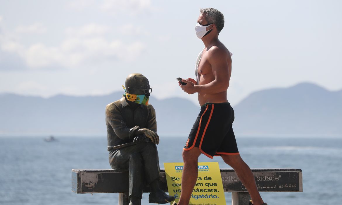 Um homem caminha ao lado da estátua do escritor brasileiro Carlos Drummond de Andrade que usa máscara protetora no primeiro dia de uso obrigatório de máscaras em meio ao surto de doença por coronavírus (COVID-19), na praia de Copacabana