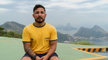Área externa. Sentado, Fabio de camisa amarela e short azul.  Ao fundo, montanhas, Baía de Guanabara e Pão de Açúcar. 