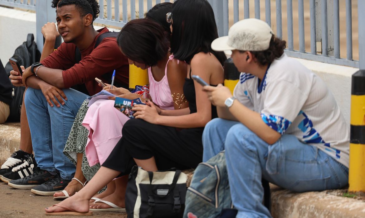 Grupo de jovens sentados na calçada, estudando e utilizando o celular antes da prova do Enem, representando a preparação dos candidatos.