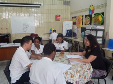 Descrição da foto: Ana Maria está sentada em uma mesa redonda, com quatro alunos, e está com os braços apoiados em um caderno aberto. Os alunos, dois meninos e duas meninas, estão sentados em semicírculo, de frente para ela, e vestem uma camisa branca.