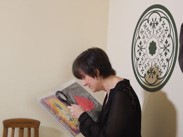 Descrição da foto: Christina Brazil está segurando um livro aberto com uma das mãos. Com a outra mão, ela está segurando uma lupa de aumento e olhando através da lente para figuras coloridas nas páginas do livro.