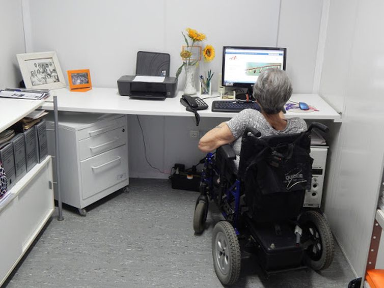 Em um escritório, a psicóloga Lilia Martins está sentada em uma cadeira de rodas motorizada, de costas para nós, e de frente para uma mesa com computador, telefone, impressora, portas-retratos e girassóis amarelos