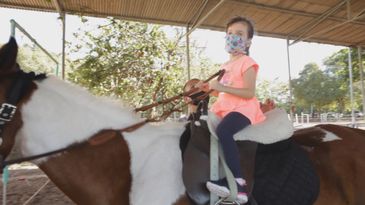 Luiza, montada em um cavalo, segura a rédea do animal.  Luiza usa uma máscara azul com desenhos coloridos.  Atrás deles, um homem de máscara preta