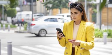 Mulher olha o celular na rua com um cartão de crédito na outra mão