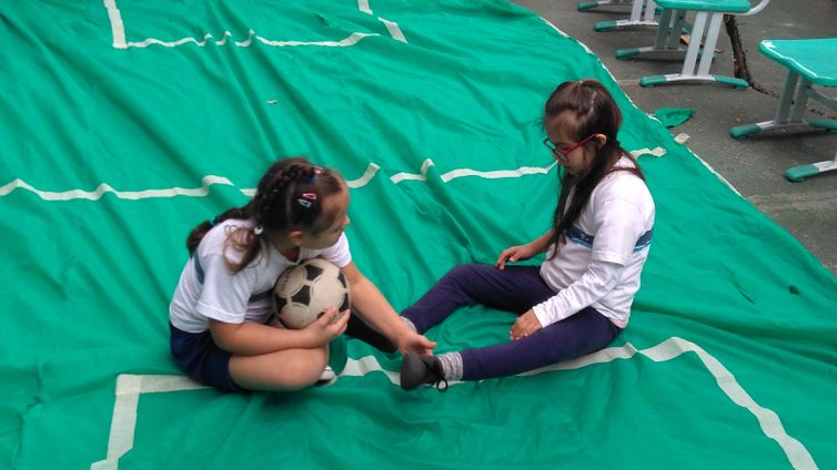 Descrição de foto: Em uma área externa, duas meninas sentadas, uma de frente para a outra, em um pano de cor verde. Uma das meninas segura uma bola.