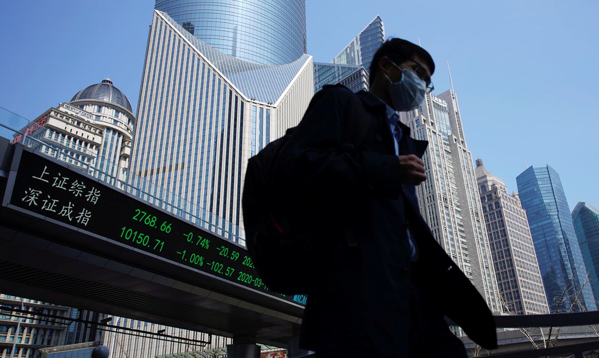 Pedestrian wearing a face mask walks near an overpass with an electronic board showing stock information in Shanghai