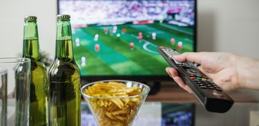 Detalhe de mão segurando controle; TV ligada no futebol; comida e bebida na mesa. 