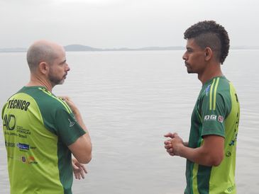 Descrição de foto: Com o mar ao fundo, Luiz Ricardo está ao lado de Eduardo Duarte. Os dois estão de frente um para o outro, de perfil, e vestem o mesmo uniforme verde.