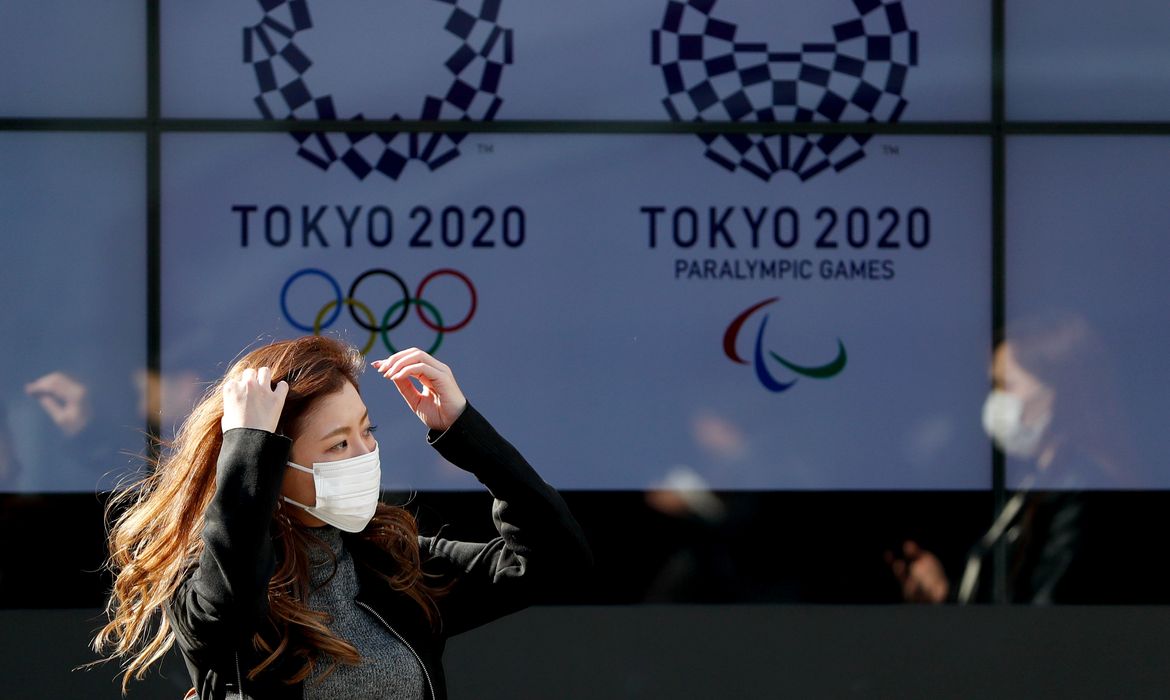 Mulher com máscara de proteção caminha em frente aos logos da Olimpíada e da Paralimpíada Tóquio-2020 / Jogos de Tóquio -anéis olimpícos, olimpíada, máscara, coi