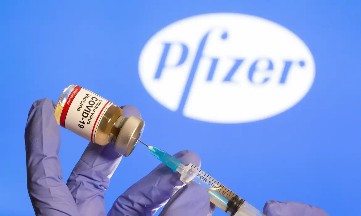 Mulher segura frasco rotulado como de vacina contra Covid-19 em frente a logo da Pfizer em foto de ilustração