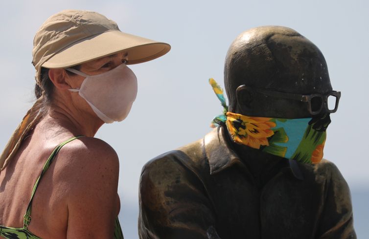 Uma mulher posa para uma foto ao lado da estátua do escritor brasileiro Carlos Drummond de Andrade que usa uma máscara protetora no primeiro dia de uso obrigatório de máscaras  em meio ao surto de doença por coronavírus (COVID-19), na praia de Copacabana