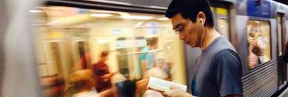 Homem lê um livro na plataforma de uma estação de metrô enquanto um trem passa