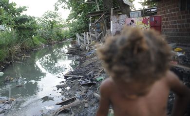 Imagem de riacho com falta de saneamento básico e uma criança passando em favela do Complexo da Maré