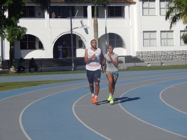 Descrição: Em uma pista de atletismo,  Diogo e Alice estão correndo juntos, lado a lado. Os dois estão com uma faixa preta de velcro envolvendo os dois braços que se encostam.