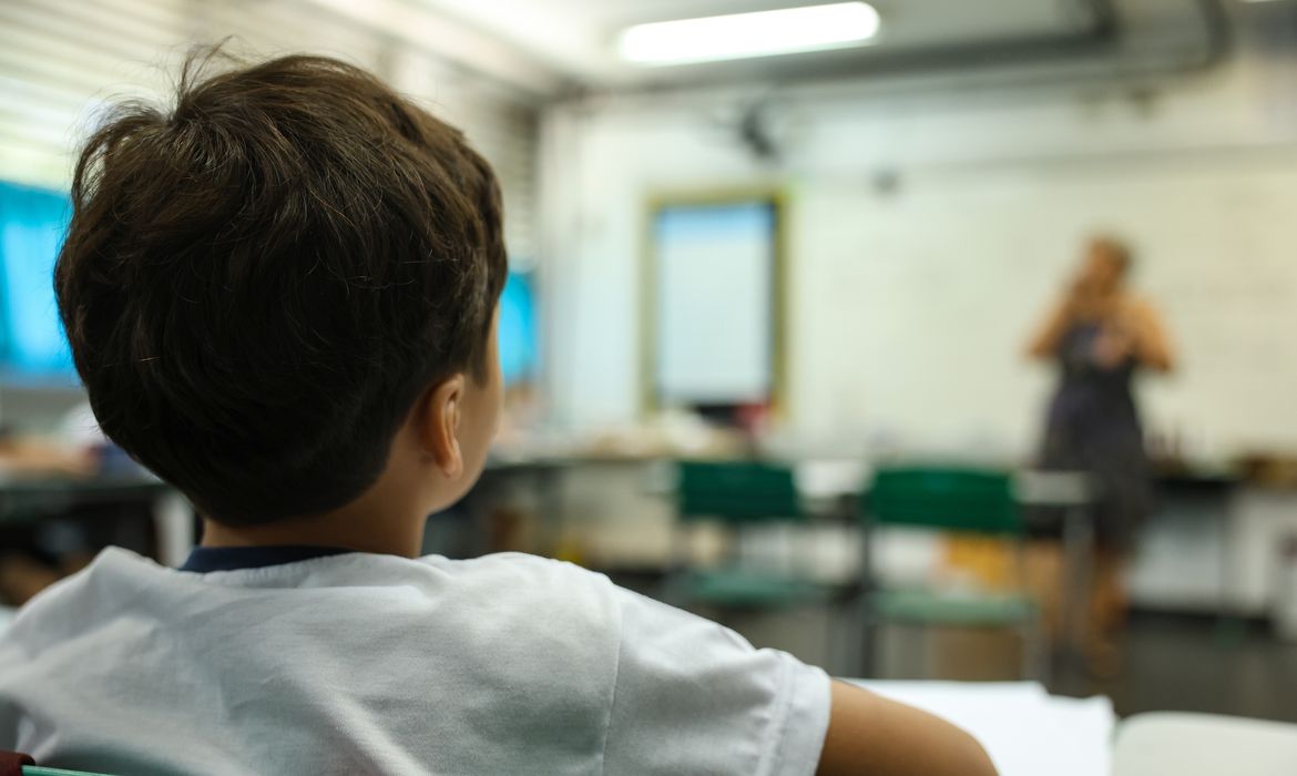Estudante em sala de aula observando a professora ao fundo, em ambiente de educação básica. Cena representa aprendizagem, ensino integral e rotina escolar.