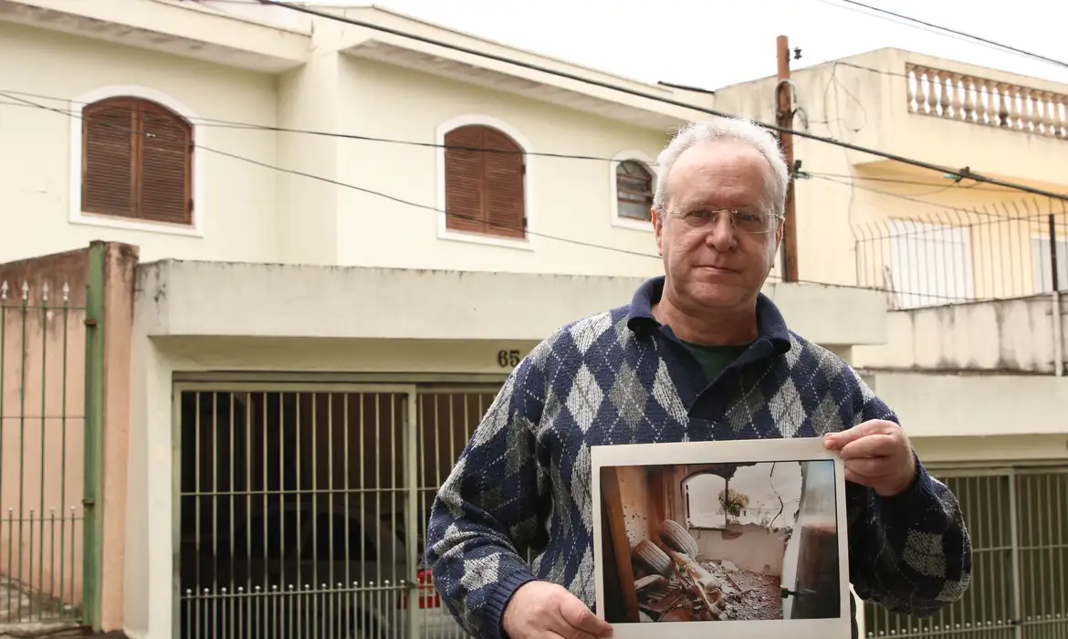 O jornalista Jorge Tadeu da Silva, um dos afetados pelo acidente com o avião modelo Fokker 100 da TAM que vitmou 99 pessoas em 1996, segura foto de como ficou a casa dele na rua Luís Orsini de Castro, no Jabaquara.