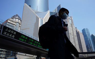 Pedestrian wearing a face mask walks near an overpass with an electronic board showing stock information in Shanghai