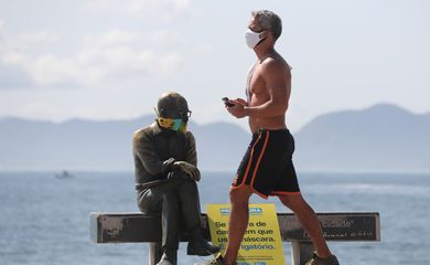 Um homem caminha ao lado da estátua do escritor brasileiro Carlos Drummond de Andrade que usa máscara protetora no primeiro dia de uso obrigatório de máscaras  em meio ao surto de doença por coronavírus (COVID-19), na praia de Copacabana