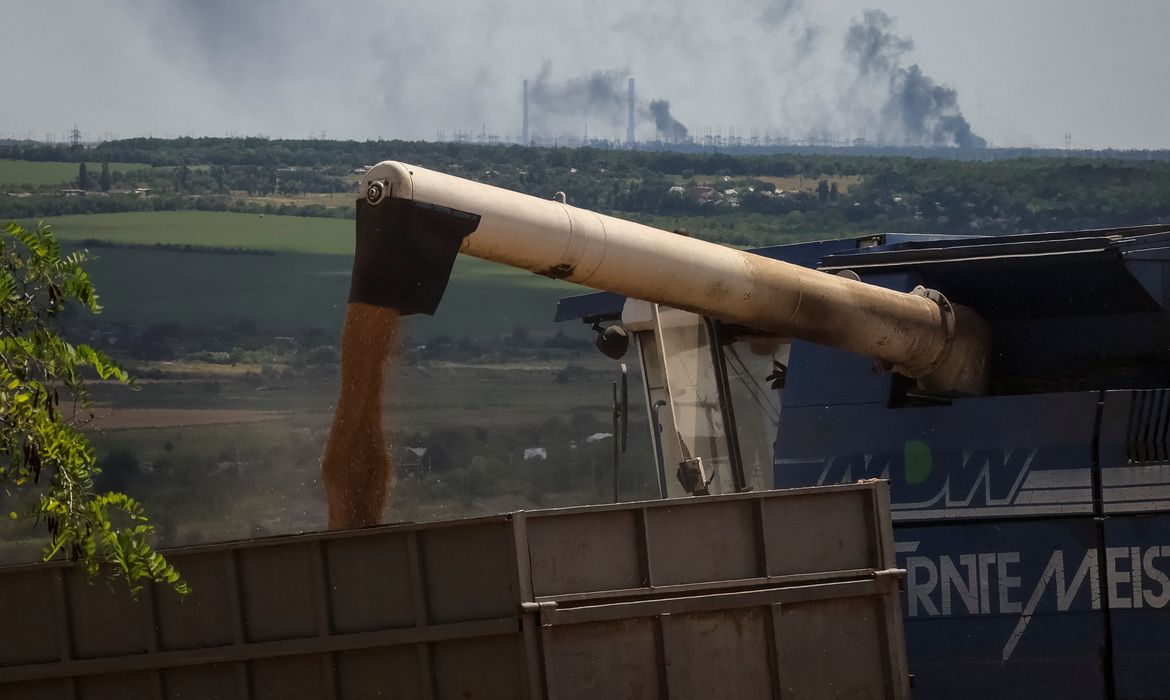 Agricultores colhem trigo na região ucraniana de Donbas enquanto usina de energia queima ao fundo, após ataque de artilharia durante invasão russa na Ucrânia