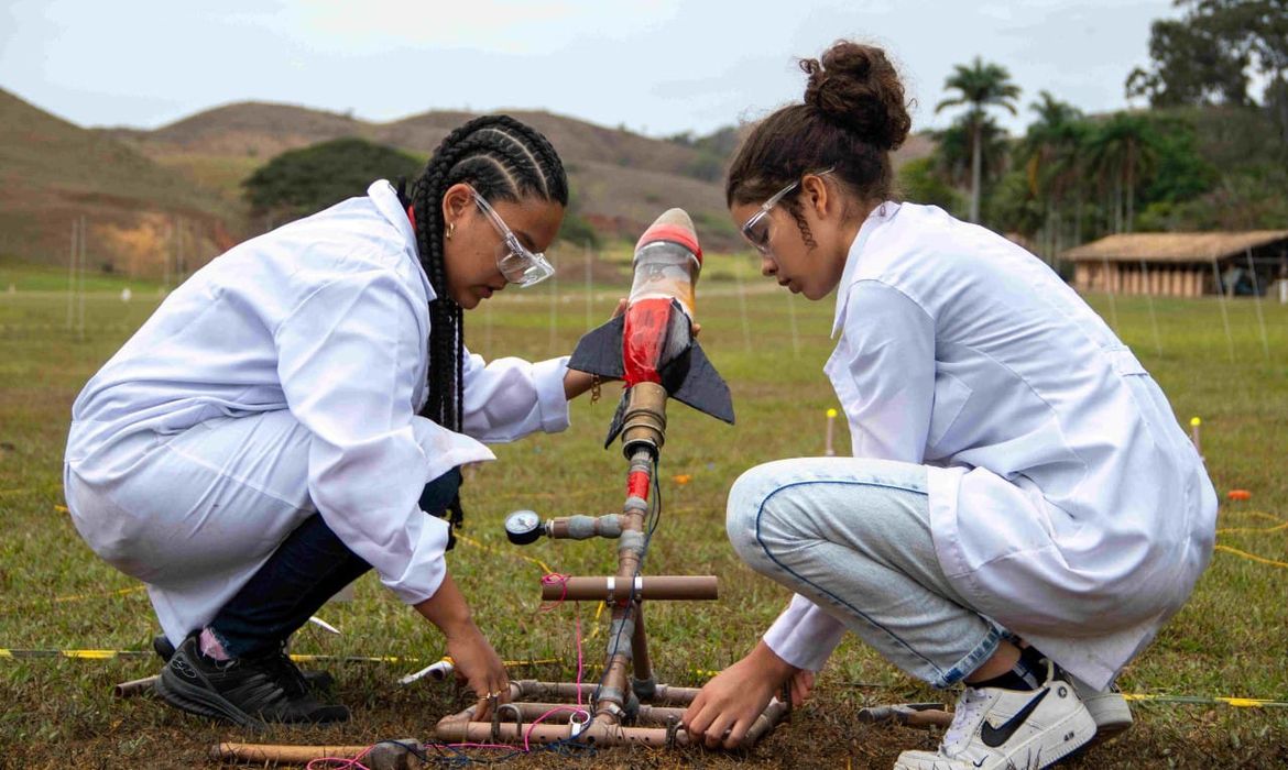 Duas estudantes usando jalecos e óculos de proteção ajustam um foguete experimental em uma base de lançamento improvisada em área aberta, durante atividade prática de ciências ao ar livre.