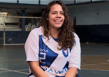 Em uma quadra, Mariana, sorridente, sentada, de blusa branca e azul da Associação de Surdos do Rio de Janeiro, ASSURJ