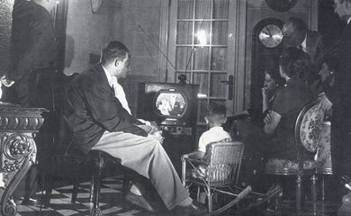 Foto em preto e branco com família em uma sala, sentada ao redor de um aparelho de televisão antigo, assiste a primeira transmissão da TV brasileira.