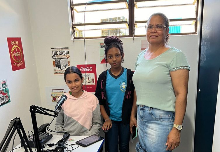 Estudantes e professora em estúdio de rádio escolar com microfone e equipamentos de gravação, atividade ligada a projetos de educação midiática nas escolas.