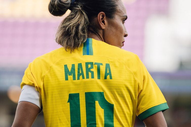 Na imagem, Marta está de costas, com o rosto virado para o lado direito, de perfil. A jogadora veste a camisa, amarela, prinicipal da Seleção, que carrega o seu nome e o número 10. No braço esquerdo, a faixa de capitã.