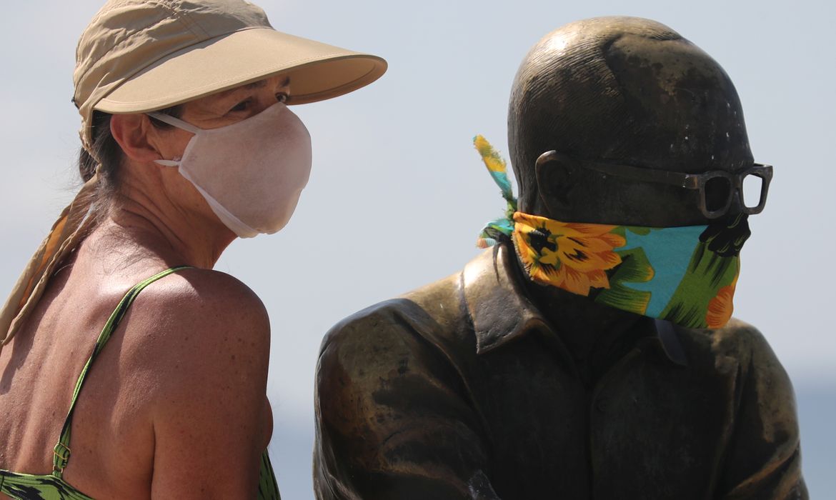 Uma mulher posa para uma foto ao lado da estátua do escritor brasileiro Carlos Drummond de Andrade que usa uma máscara protetora no primeiro dia de uso obrigatório de máscaras  em meio ao surto de doença por coronavírus (COVID-19), na praia de Copacabana