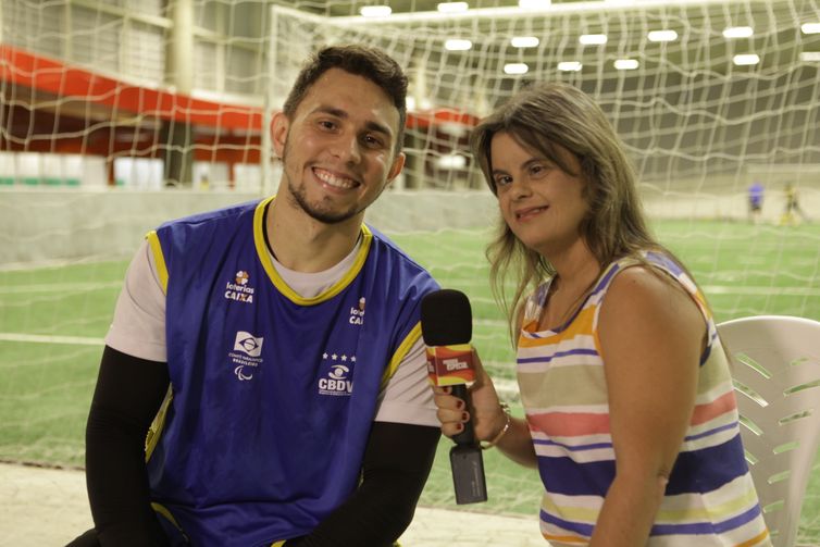  Sentados lado a lado, de costas para um gol e um campo, Luan Gonçalves e Fernanda Honorato, com o microfone do Programa Especial na mão direita