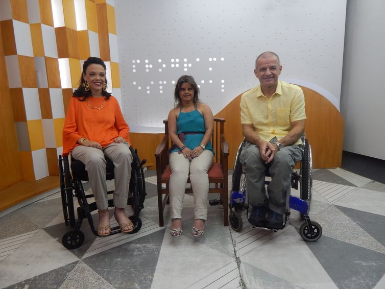 Descrição da foto: No estúdio do Programa Especial, Juliana Oliveira sentada na cadeira de rodas. Ao lado dela, Fernanda Honorato sentada em uma cadeira de madeira, e Zé Luiz Pacheco sentado em uma cadeira de rodas.