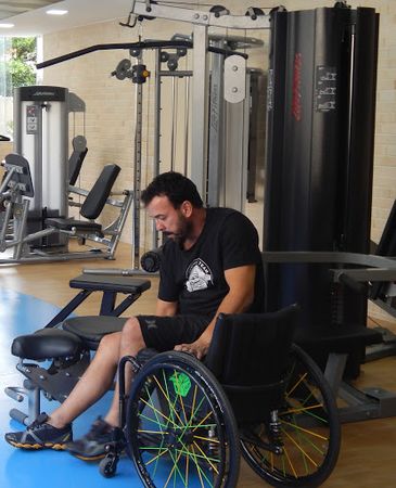 Descrição da foto: Em uma academia de ginástica, Eduardo Mayr senta em um dos aparelhos, apoiando-se com uma das mãos na sua cadeira de rodas, que está virada para ele.