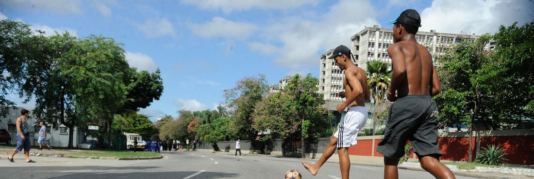 Meninos da comunidade Metrô-Mangueira jogam futebol e interagem com turistas em rua interditada por policiais nos arredores do estádio do Maracanã.