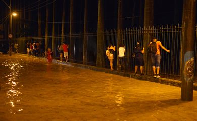 Moradores caminham segurando uma cerca ao lado de uma rua alagada durante fortes chuvas no bairro Jardim Botânico