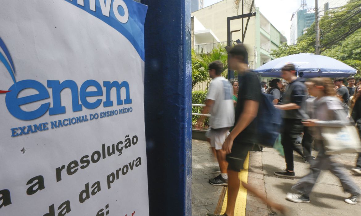 Estudantes caminham em direção ao local de aplicação do Enem. À esquerda, um cartaz com o logotipo do Exame Nacional do Ensino Médio indica informações sobre a prova.