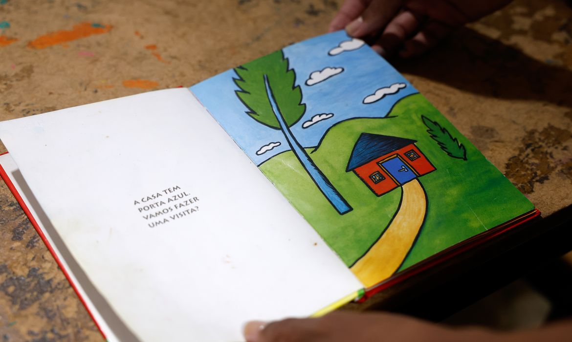 Livro infantil aberto sobre uma mesa mostra ilustração colorida de uma casa com porta azul e árvore ao lado. Na página ao lado, lê-se o texto “A casa tem porta azul. Vamos fazer uma visita?