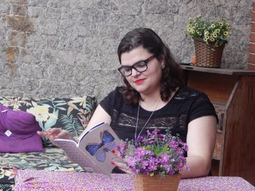 Sentada, Emiliana Rosa segura o livro &quot;Borboletas Poéticas&quot; com as duas mãos. Ela usa óculos de grau.